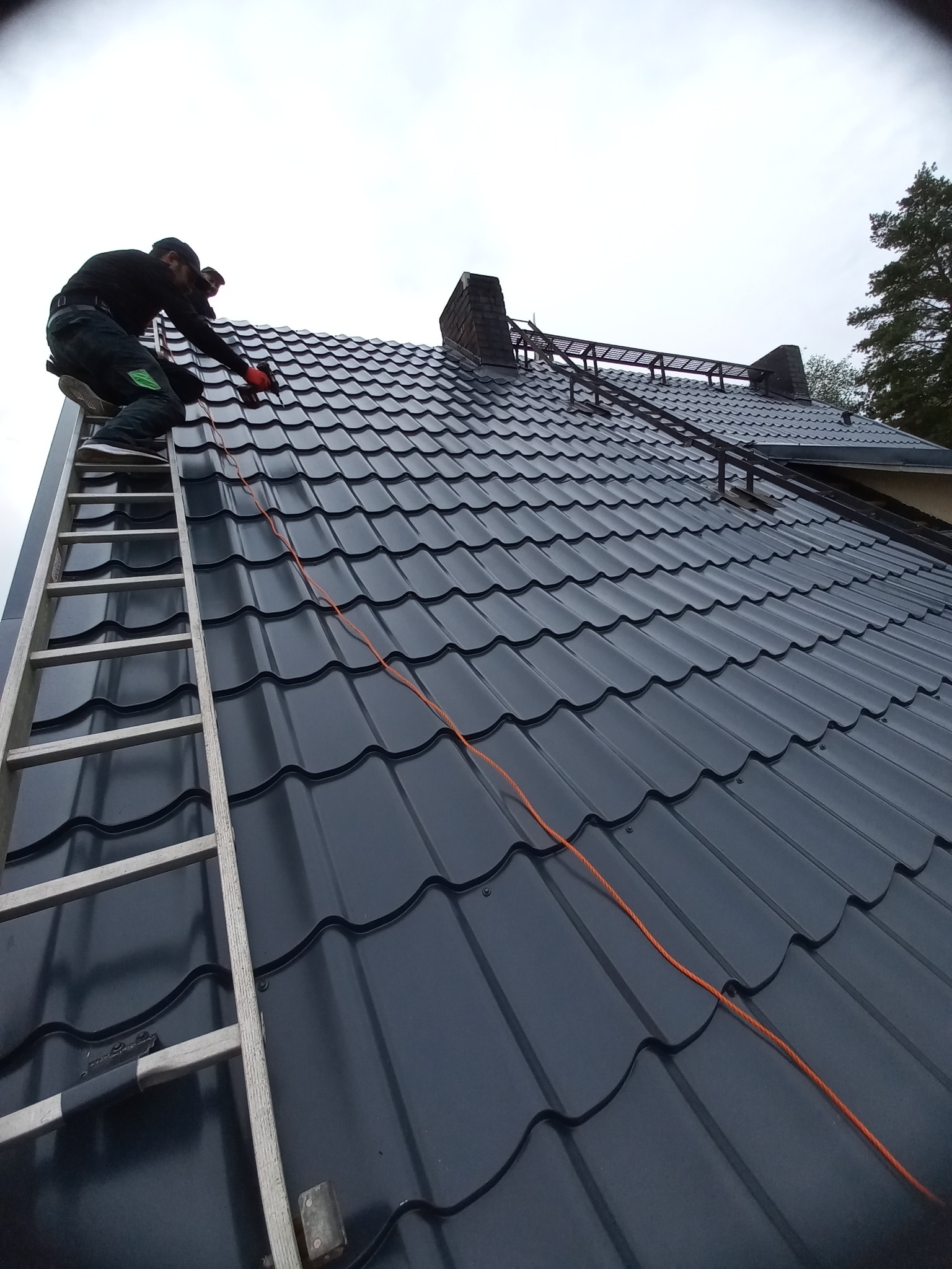 Pracownik w rękawicach na drabinie montuje ciemną blachę na dachu domu, widoczny komin i metalowa konstrukcja zabezpieczająca.