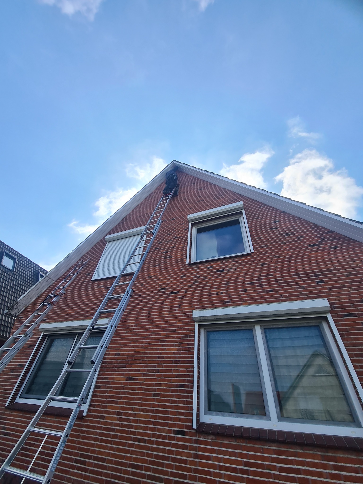 Pracownik na drabinie przy ceglanym domu z oknami z roletami. Ujęcie z dołu, widoczne niebieskie niebo z chmurami. Druga drabina oparta o ścianę.