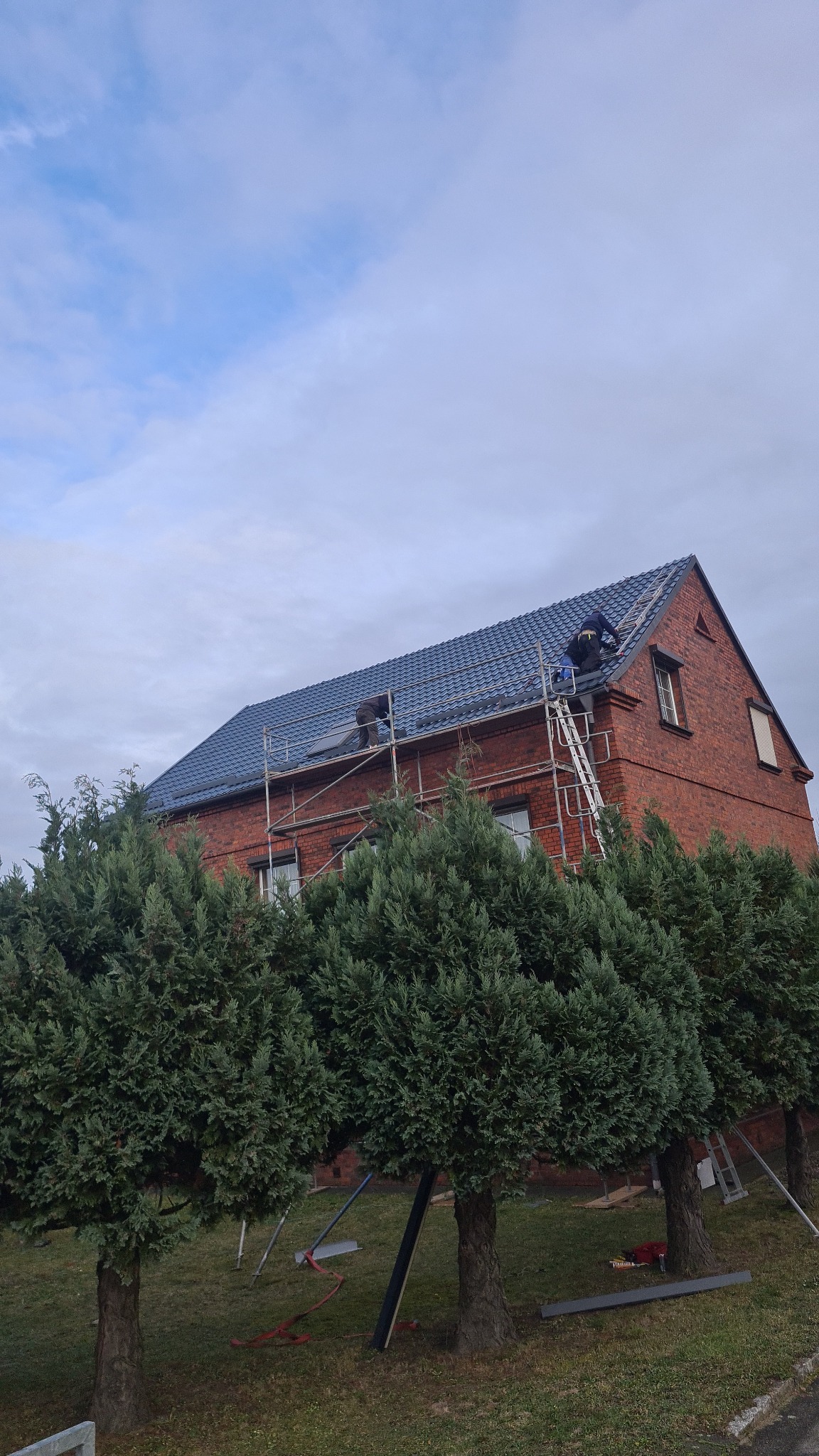 Ceglany dom z nowym, niebieskim dachem krytym dachówką. Na dachu pracownicy na rusztowaniu i drabinie. Część dachu jeszcze niepokryta. W tle pochmurne niebo.