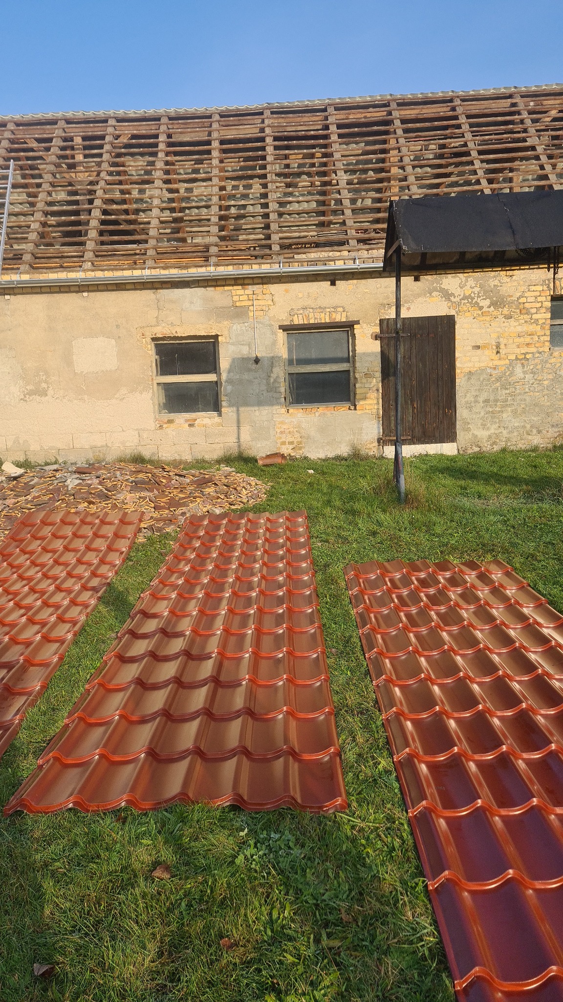 Ceglasta blacha dachowa na trawie przed budynkiem z odsłoniętą więźbą, ukazująca etap prac dekarskich przy wymianie pokrycia dachowego.