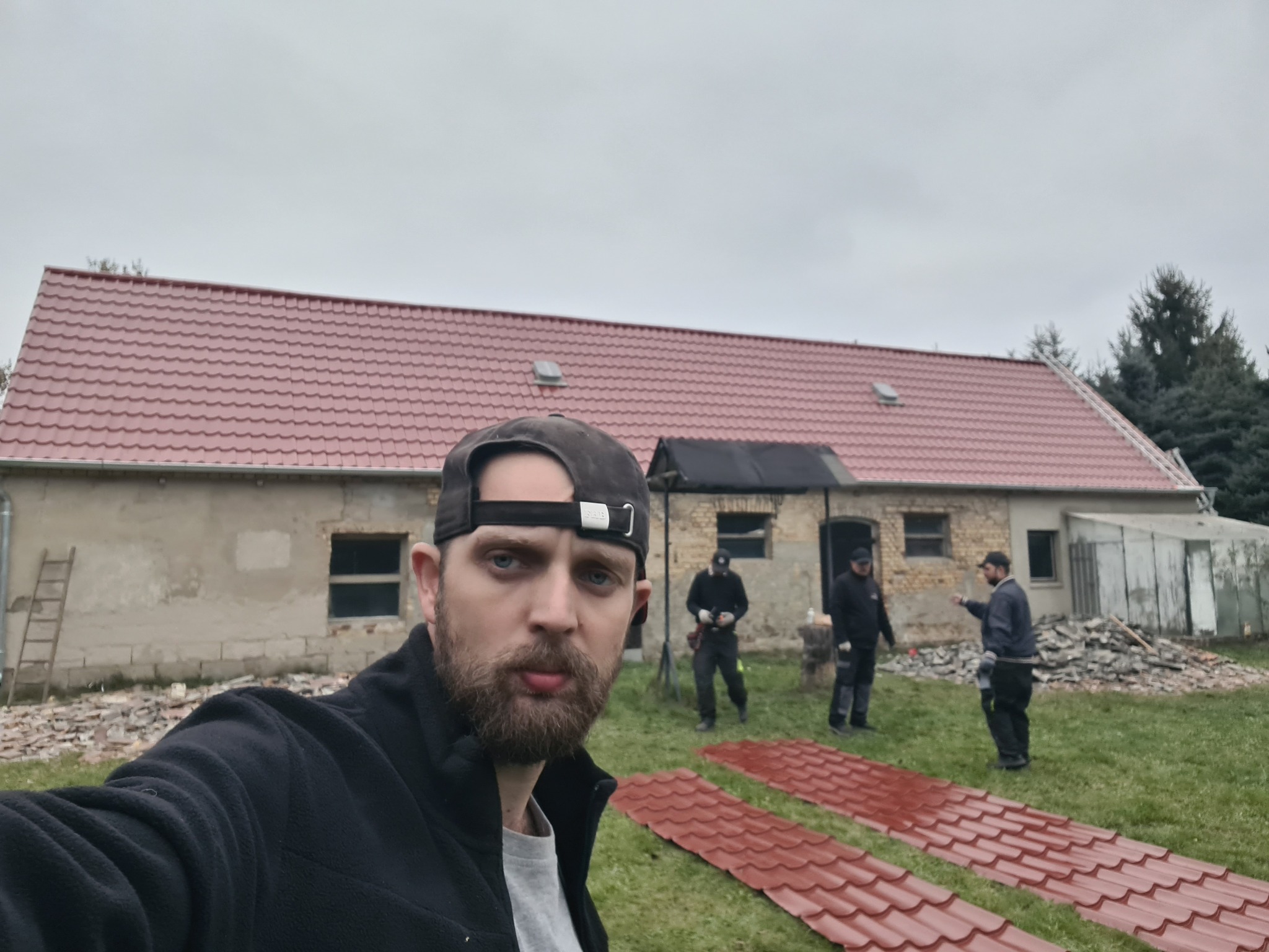 Mężczyzna robi selfie na tle domu z nowym, czerwonym dachem. W tle ekipa montuje dachówkę. Na trawie leżą przygotowane elementy pokrycia.