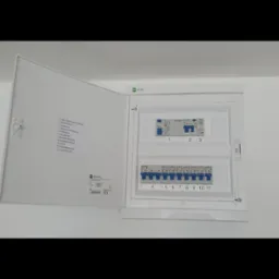 Otwarta skrzynka rozdzielcza z widocznymi bezpiecznikami i listą obwodów, zamontowana na białej ścianie w nowym budownictwie.