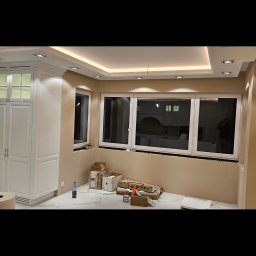 SZATER MATEUSZ BAR - Wnętrze w trakcie wykańczania: białe szafki kuchenne z oświetleniem LED, marmurowa podłoga, kartony i butelka wody mineralnej przy ścianie z trzema oknami, widoczne punkty świetlne w suficie.