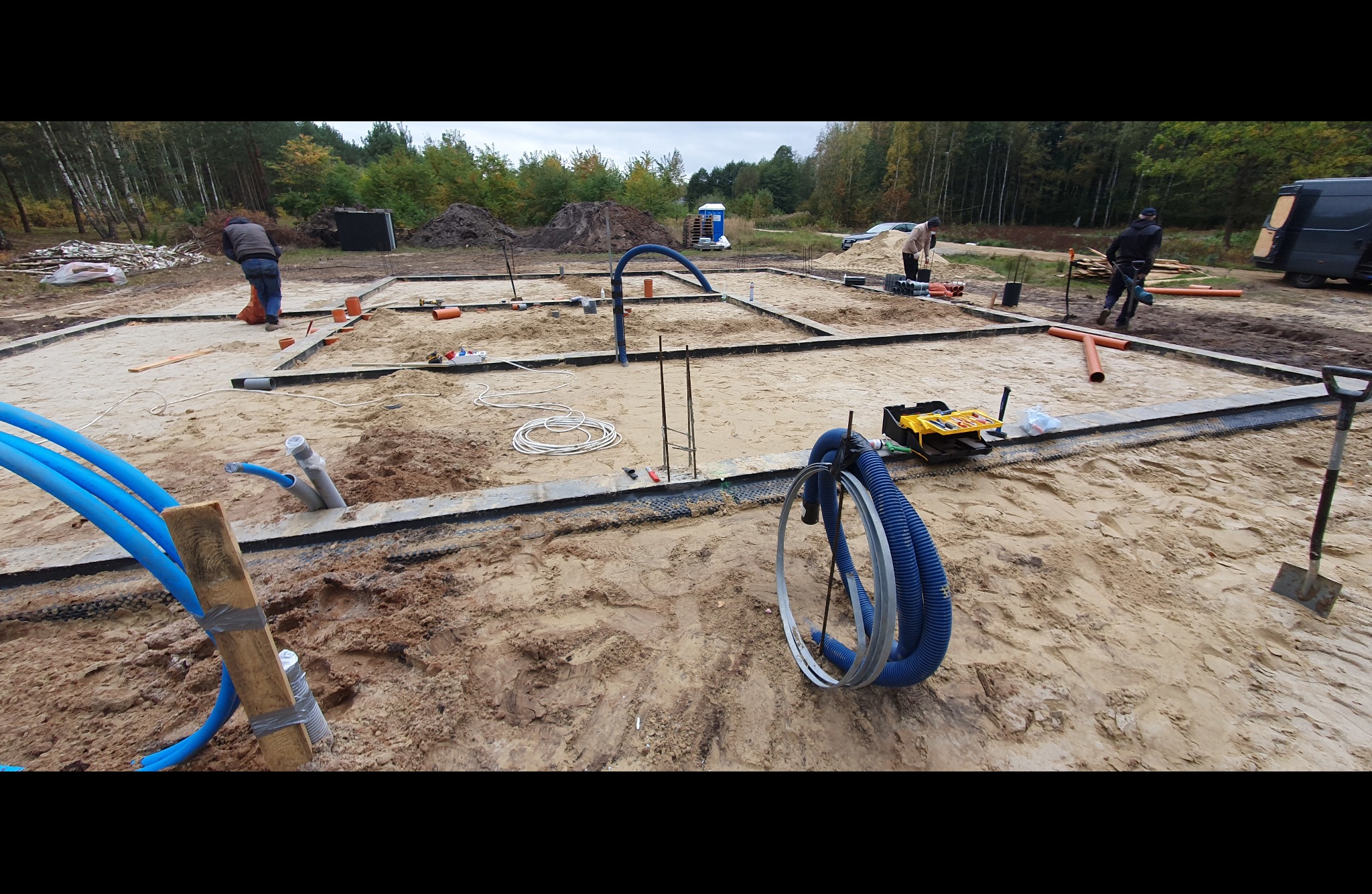 Przygotowanie terenu pod fundamenty budynku: wykopane rowy, ułożone szalunki, wystające pręty zbrojeniowe, widoczne rury instalacyjne, narzędzia i pracownicy w tle.