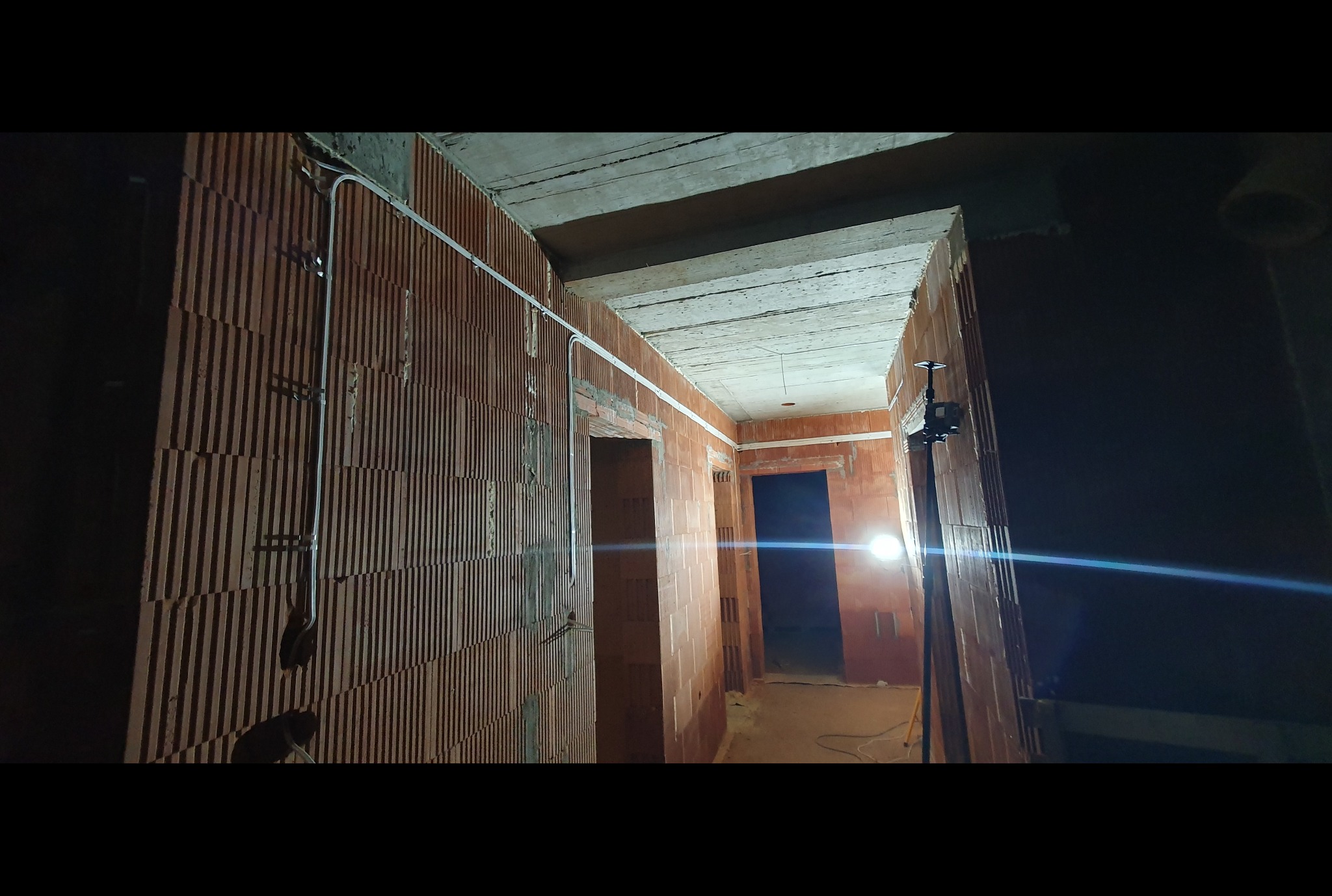 Wnętrze w trakcie budowy z widocznymi przewodami elektrycznymi zamocowanymi na ścianie z cegły dziurawki, oświetlone lampą budowlaną na statywie.