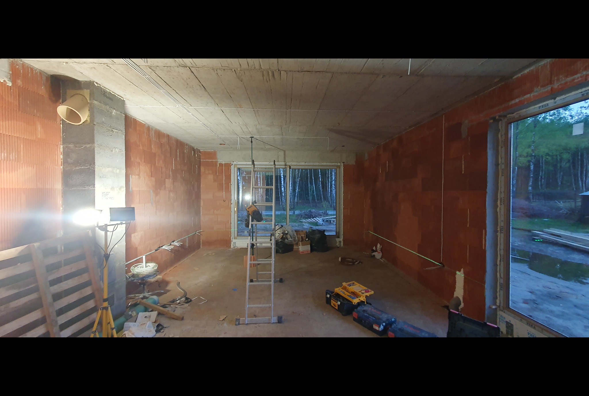 Wnętrze w stanie surowym z widocznymi przewodami elektrycznymi na suficie, drabina aluminiowa, narzędzia i lampa budowlana. Ściany z cegły dziurawki, surowy betonowy strop.