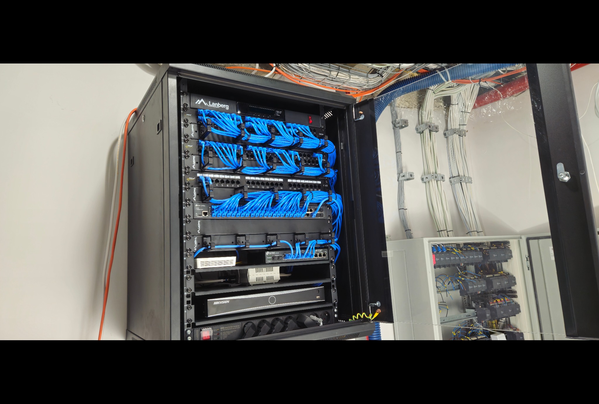 Otwarta szafa rack Lanberg z panelami krosowniczymi i niebieskimi kablami patchcord, obok widoczna skrzynka rozdzielcza z zabezpieczeniami, kable prowadzone w korytach pod sufitem.