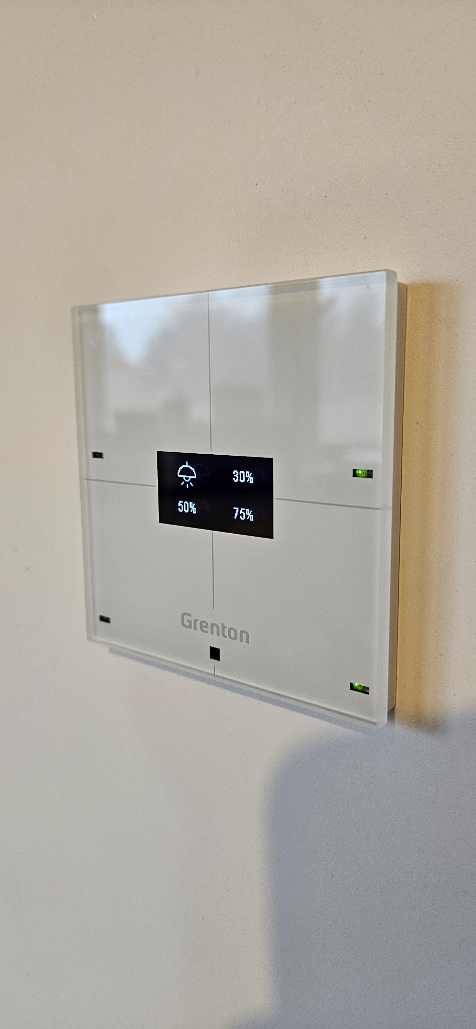 Panel sterowania inteligentnego domu marki Grenton, zamontowany na ścianie, wyświetlający informacje o wilgotności i natężeniu światła.