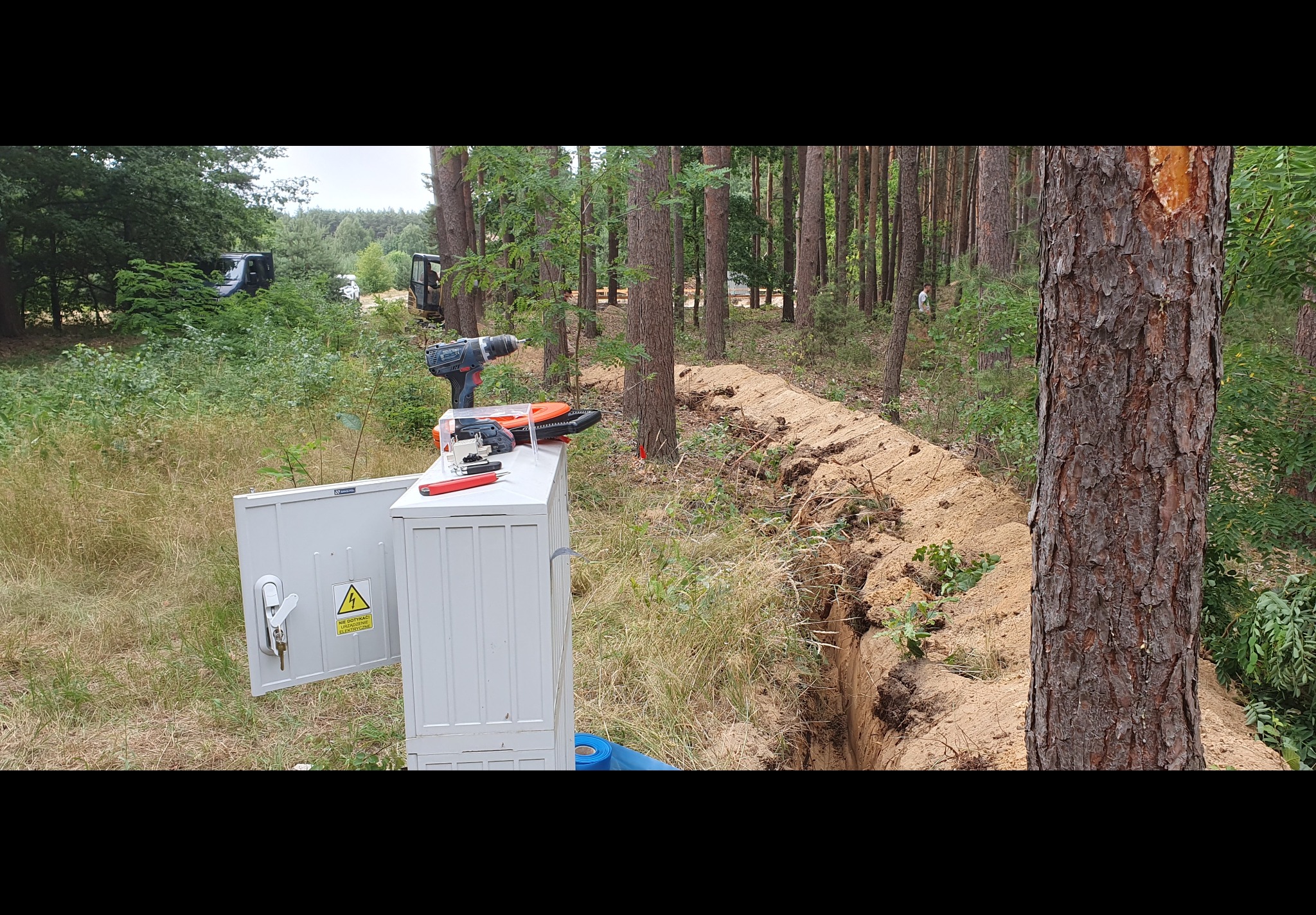 Otwarta skrzynka elektryczna z narzędziami na pokrywie, obok wykopany rów w zalesionym terenie; w tle widoczny sprzęt budowlany.