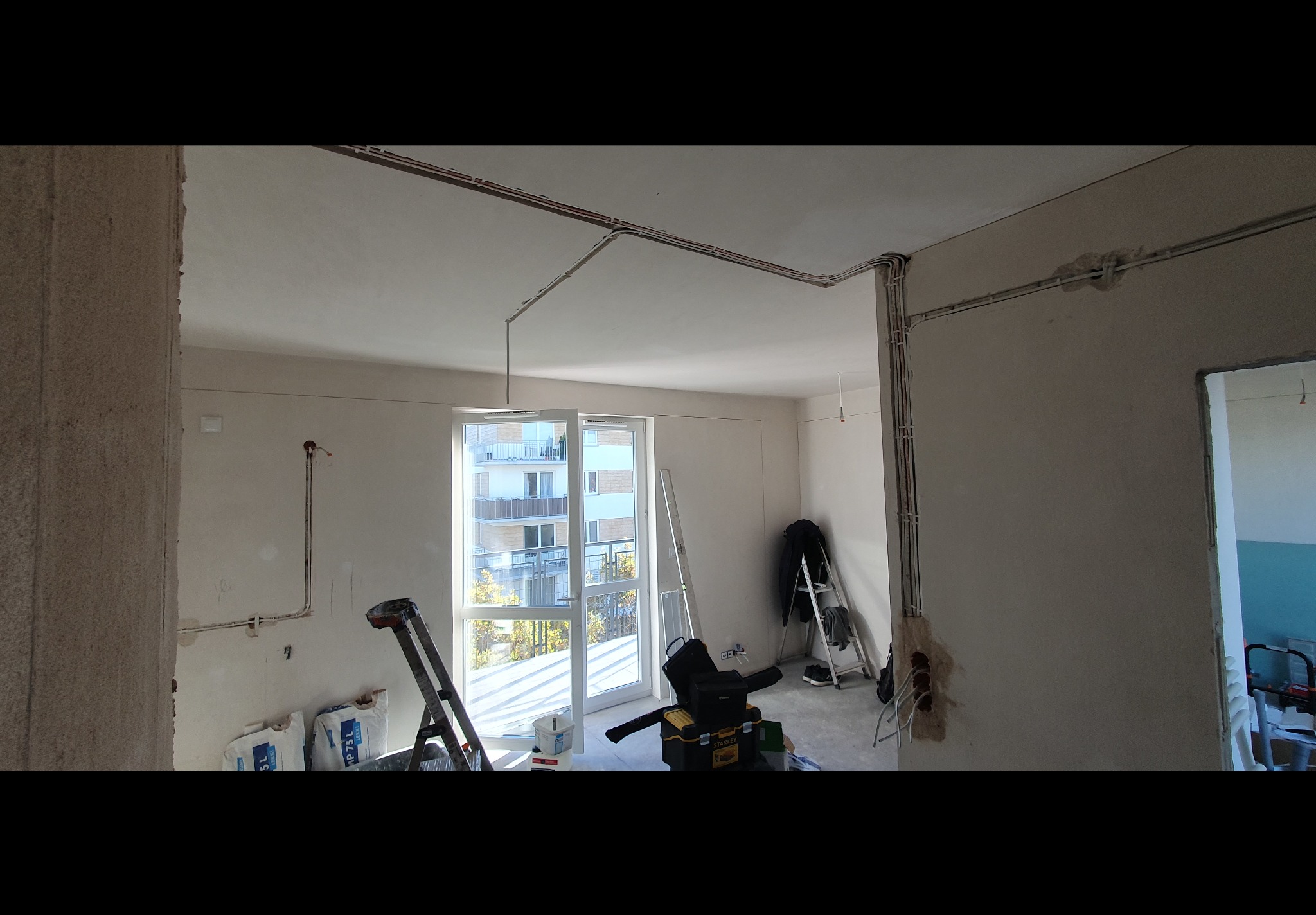 Wnętrze w stanie surowym z widocznymi przewodami elektrycznymi prowadzonymi po ścianach i suficie, drabiny, torby z zaprawą i narzędzia sugerują prace wykończeniowe.