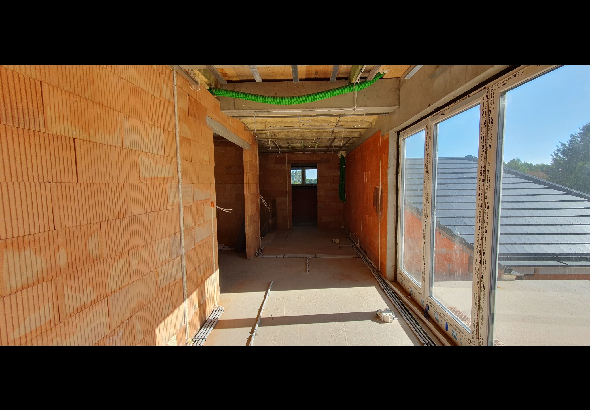 Wnętrze w stanie surowym otwartym, widoczne pomarańczowe bloczki ceramiczne, rozprowadzone instalacje elektryczne w szarych rurkach, zielona rura karbowana pod sufitem i okna z widokiem na dach.