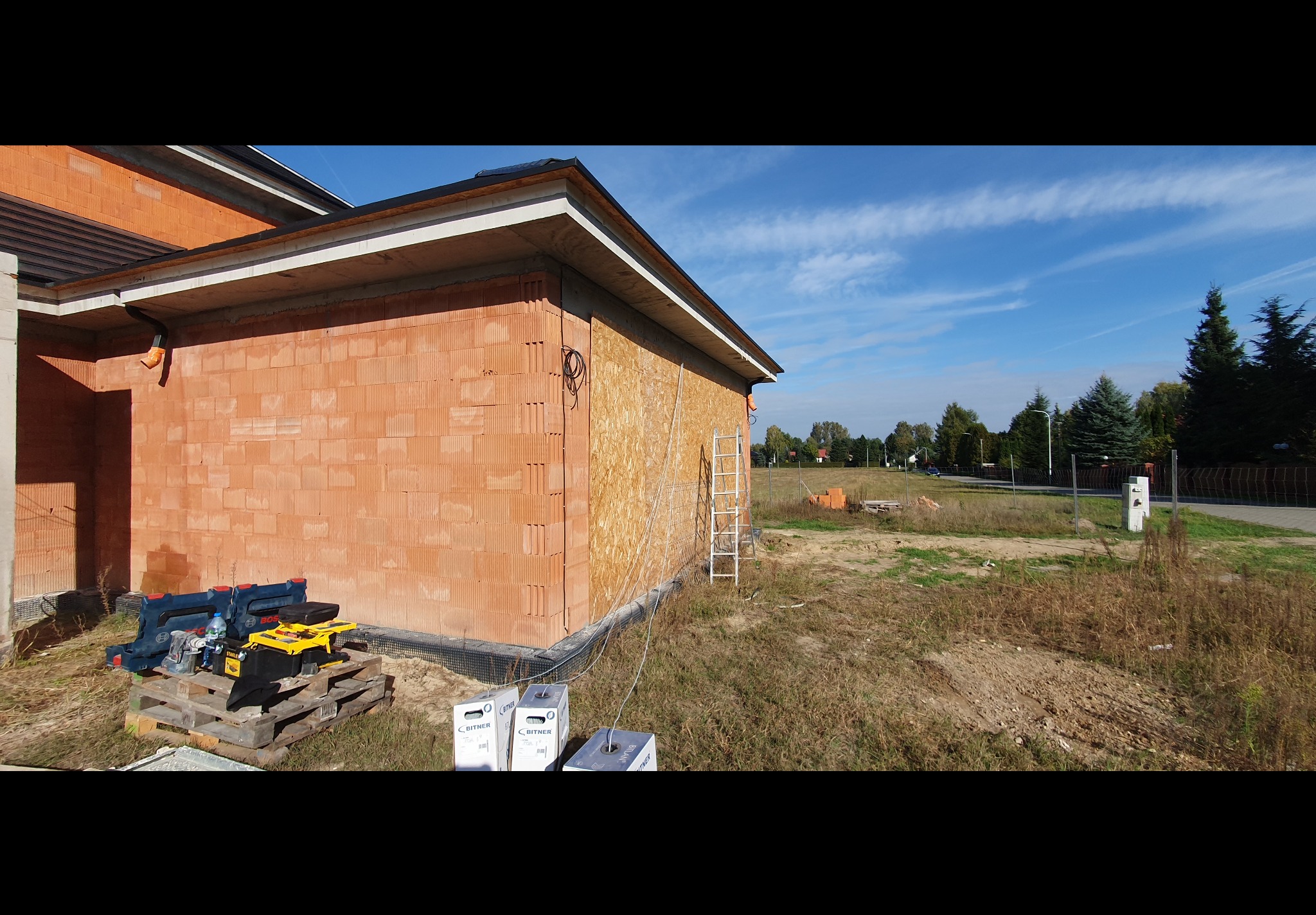 Budynek w trakcie budowy z widocznym ociepleniem ścian, narzędziami na palecie i zwiniętymi kablami na zewnątrz, na tle błękitnego nieba.