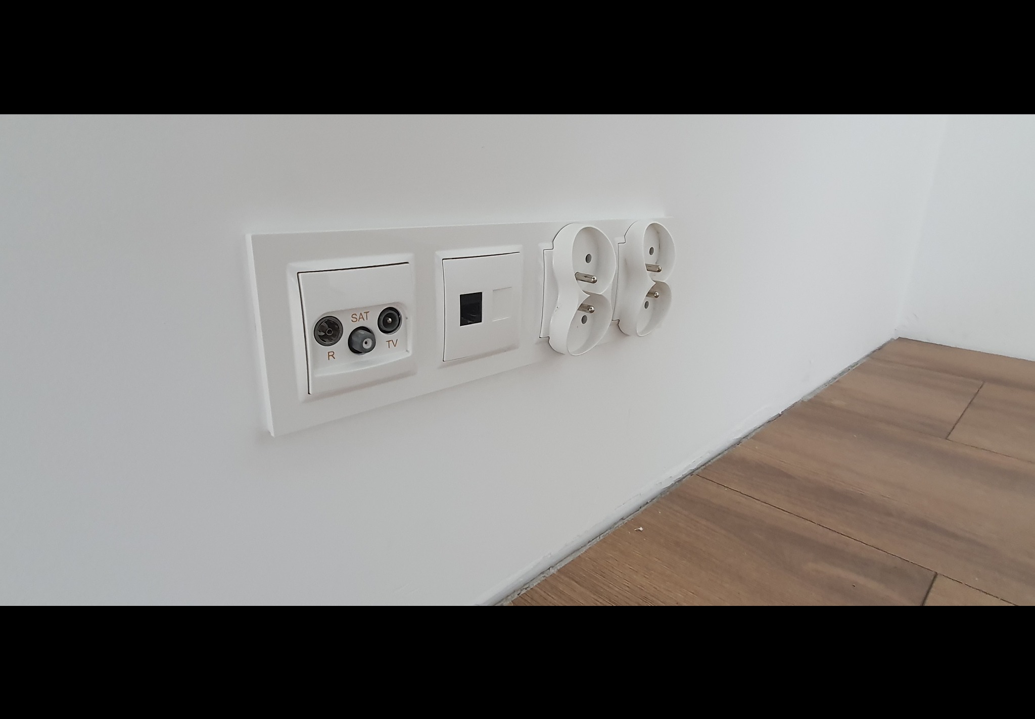 Białe gniazdka elektryczne, gniazdo antenowe SAT/TV oraz gniazdo internetowe zamontowane na białej ścianie, widoczny fragment podłogi imitującej drewno.