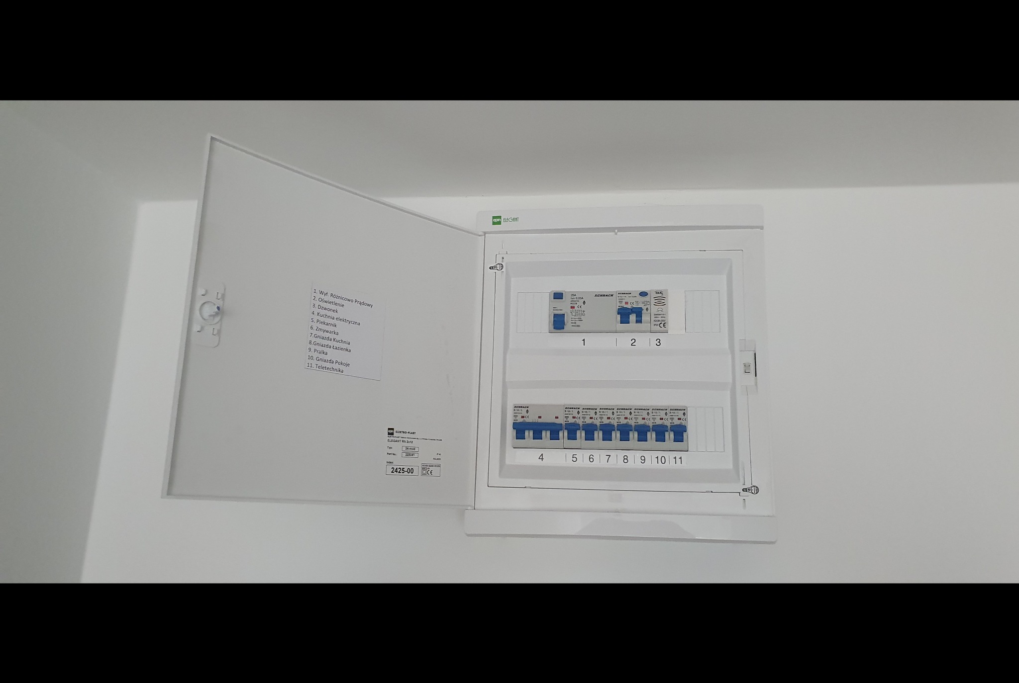 Otwarta skrzynka rozdzielcza z widocznymi bezpiecznikami i listą obwodów, zamontowana na białej ścianie w nowym budownictwie.