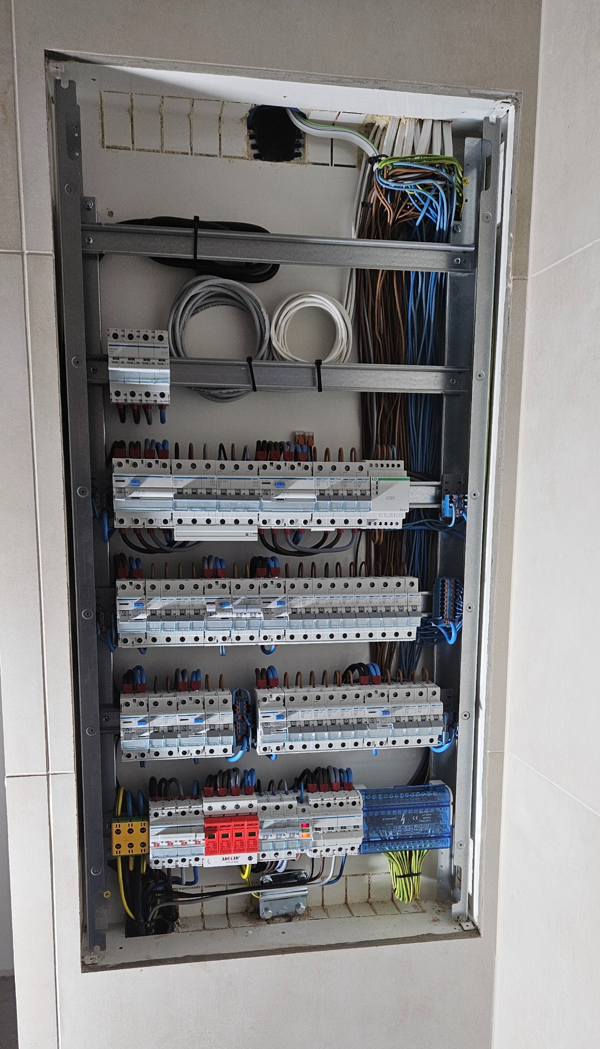 Otwarta szafa rozdzielcza z licznymi bezpiecznikami, przewodami w różnych kolorach i listwami zaciskowymi, zamontowana w ścianie wykończonej płytkami.