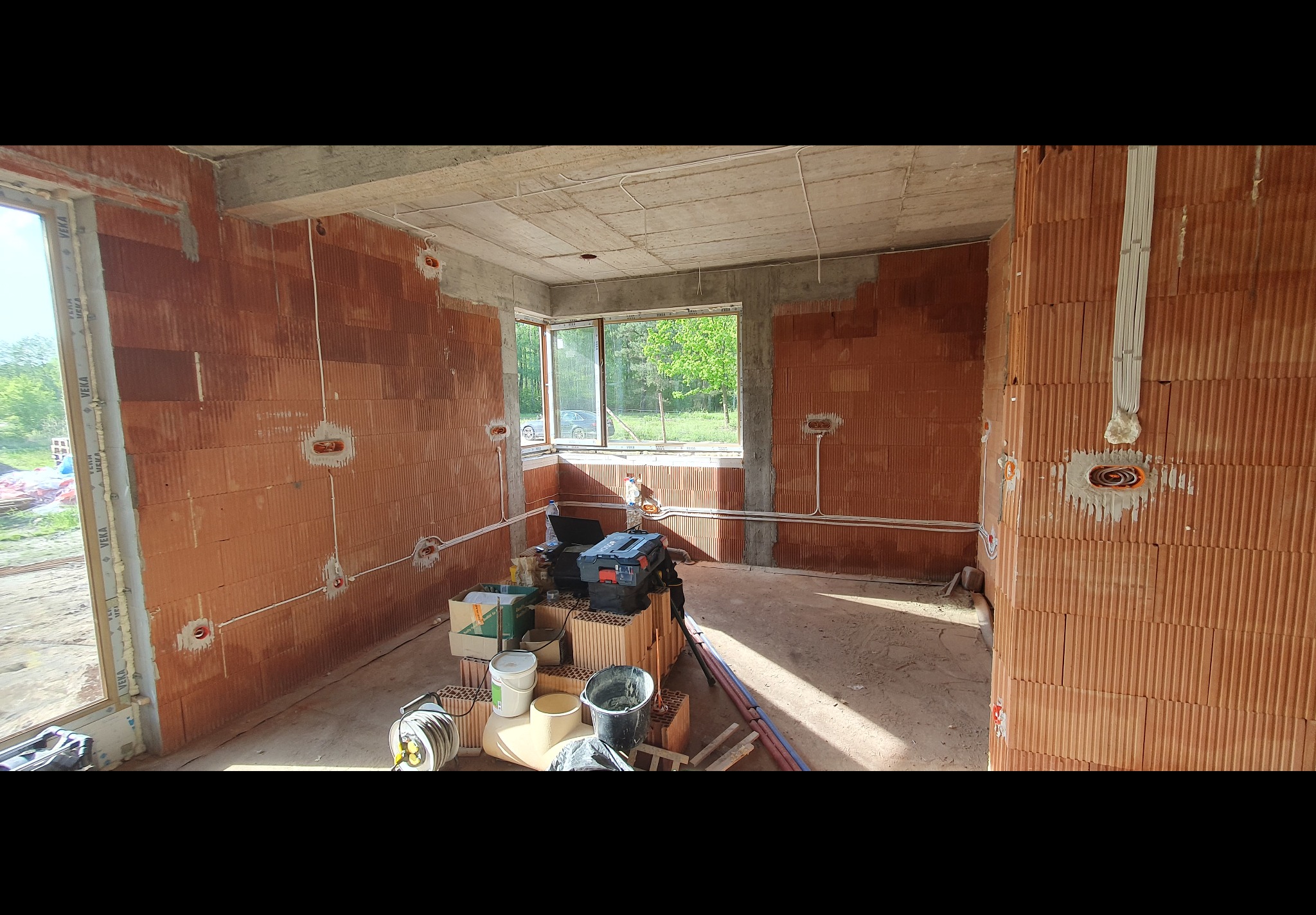 Wnętrze surowego domu w trakcie montażu instalacji elektrycznej: widoczne przewody poprowadzone po ścianach z czerwonej cegły, puszki elektryczne, okno z widokiem na zieleń oraz narzędzia...