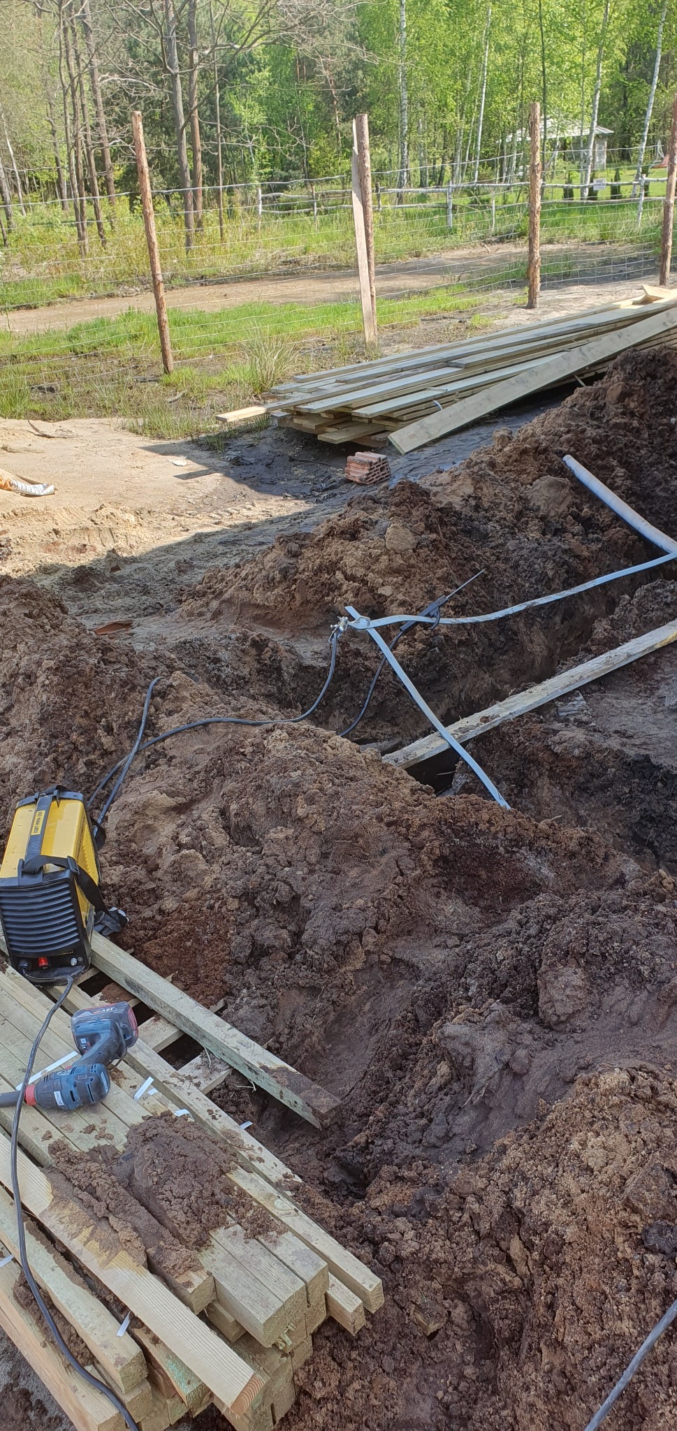 Wykop w ziemi z widocznymi kablami, spawarka i wkrętarki leżące na drewnianych belkach, przygotowania do prac ziemnych na zewnątrz.