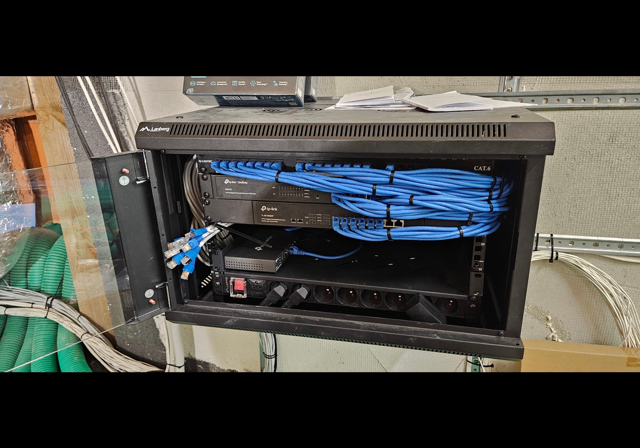 Otwarta szafa rack z zainstalowanymi urządzeniami sieciowymi, w tym routerami TP-Link i kablami CAT6 w kolorze niebieskim, ułożonymi w schludne wiązki. Widoczne gniazda zasilające i kable...