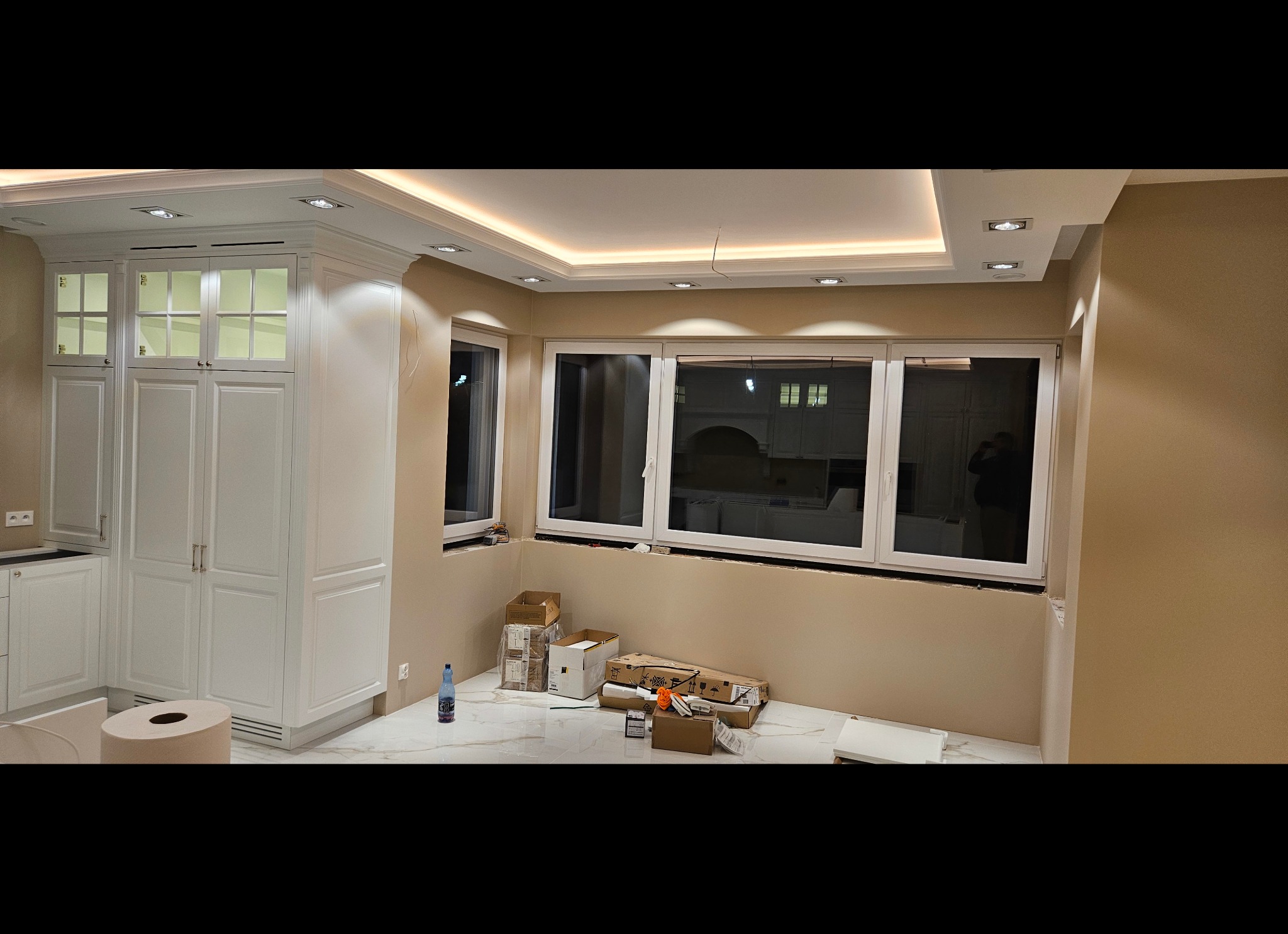 Wnętrze w trakcie wykańczania: białe szafki kuchenne z oświetleniem LED, marmurowa podłoga, kartony i butelka wody mineralnej przy ścianie z trzema oknami, widoczne punkty świetlne w suficie.