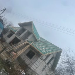 KaroDACH Dawid Sosiński - Surowa konstrukcja domu w trakcie budowy z widocznym szkieletem dachu pokrytym zieloną membraną, drewniane łaty przygotowane do dalszych prac, pochmurne niebo w tle.