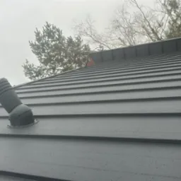 Szary dach z blachy na domu w Sterdyniu z kominem i wentylacją dachową, widoczna drabina dachowa. Widok w pochmurny dzień.