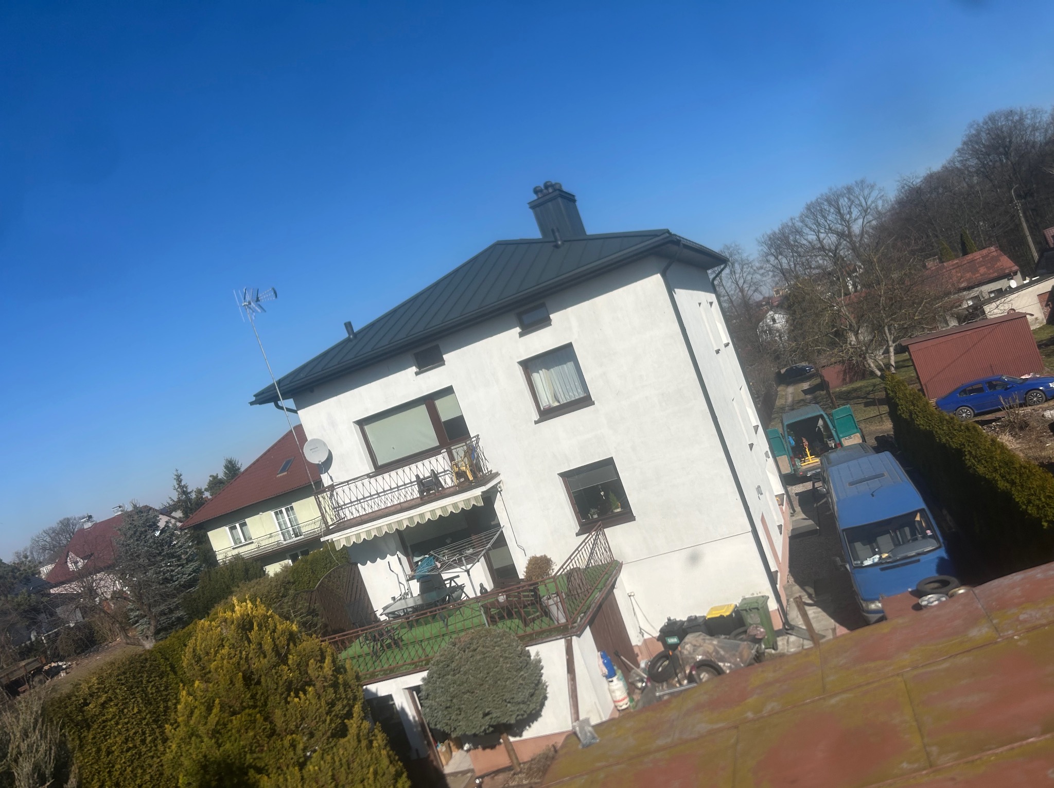 Dwupiętrowy dom z ciemnoszarym dachem w słoneczny dzień, widok z góry, w tle drzewa i inne budynki.