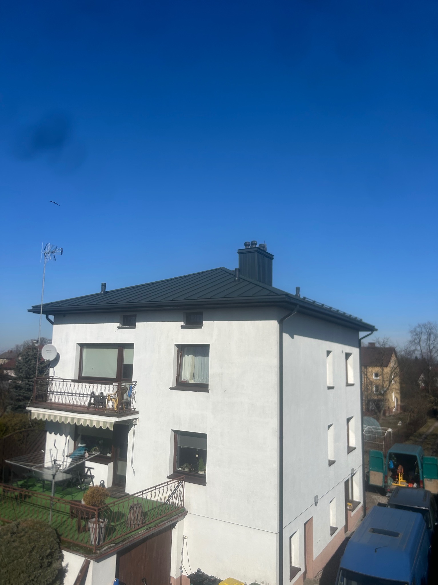 Dwupiętrowy dom z ciemnoszarym dachem, widok z góry, błękitne niebo, anteny na dachu, samochody dostawcze w tle.