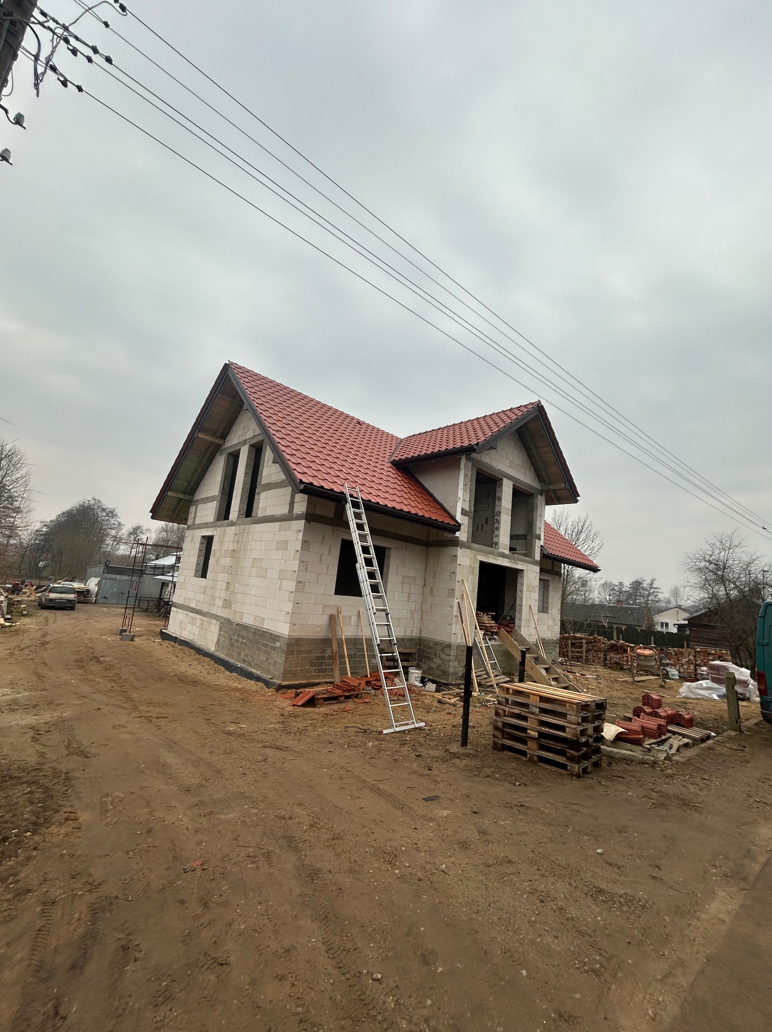 Surowy stan domu jednorodzinnego z czerwonym dachem, widoczne rusztowania, drabina oparta o ścianę, materiały budowlane na ziemi, pochmurne niebo w tle.