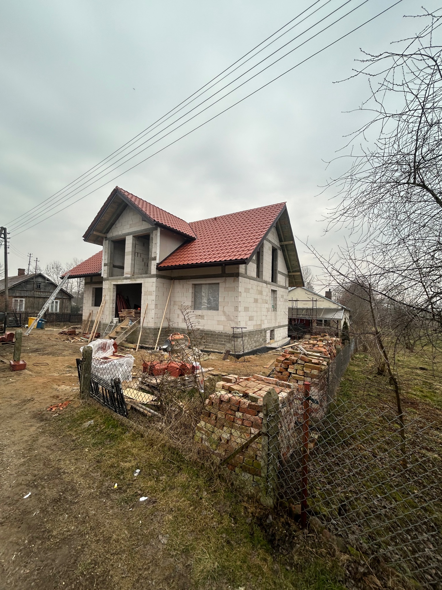 Dom w trakcie budowy z czerwonym dachem, widoczne pustaki, cegły i drabina, pochmurne niebo.