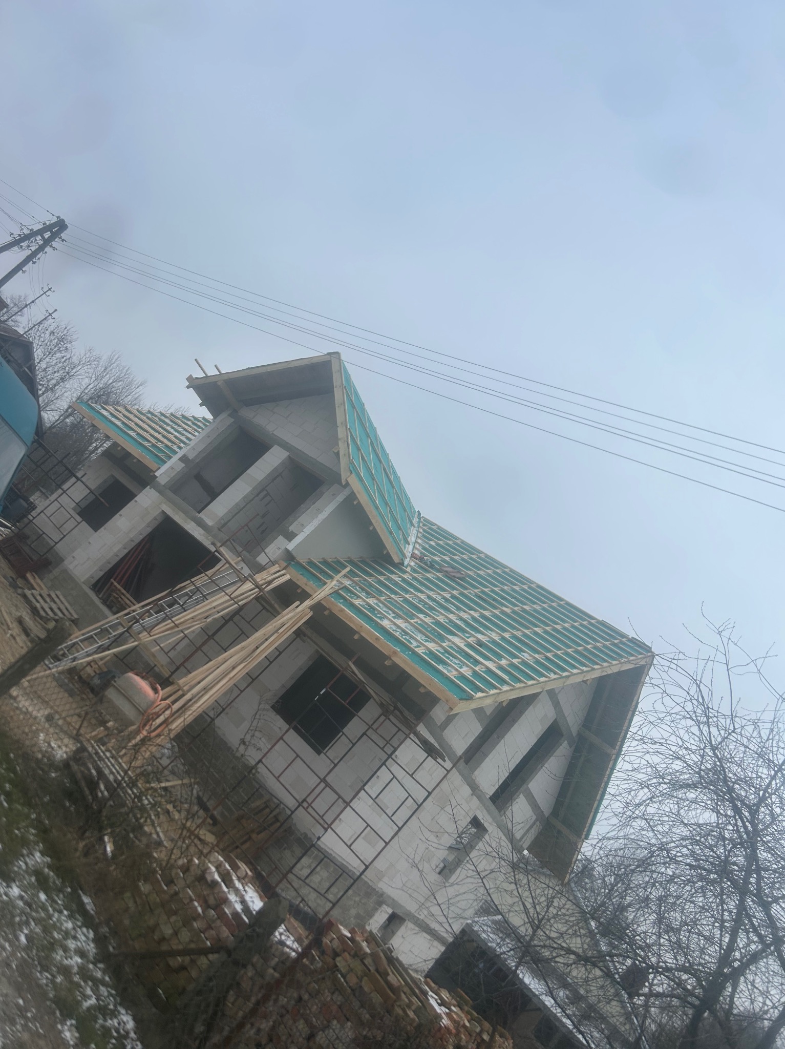 Surowa konstrukcja domu w trakcie budowy z widocznym szkieletem dachu pokrytym zieloną membraną, drewniane łaty przygotowane do dalszych prac, pochmurne niebo w tle.