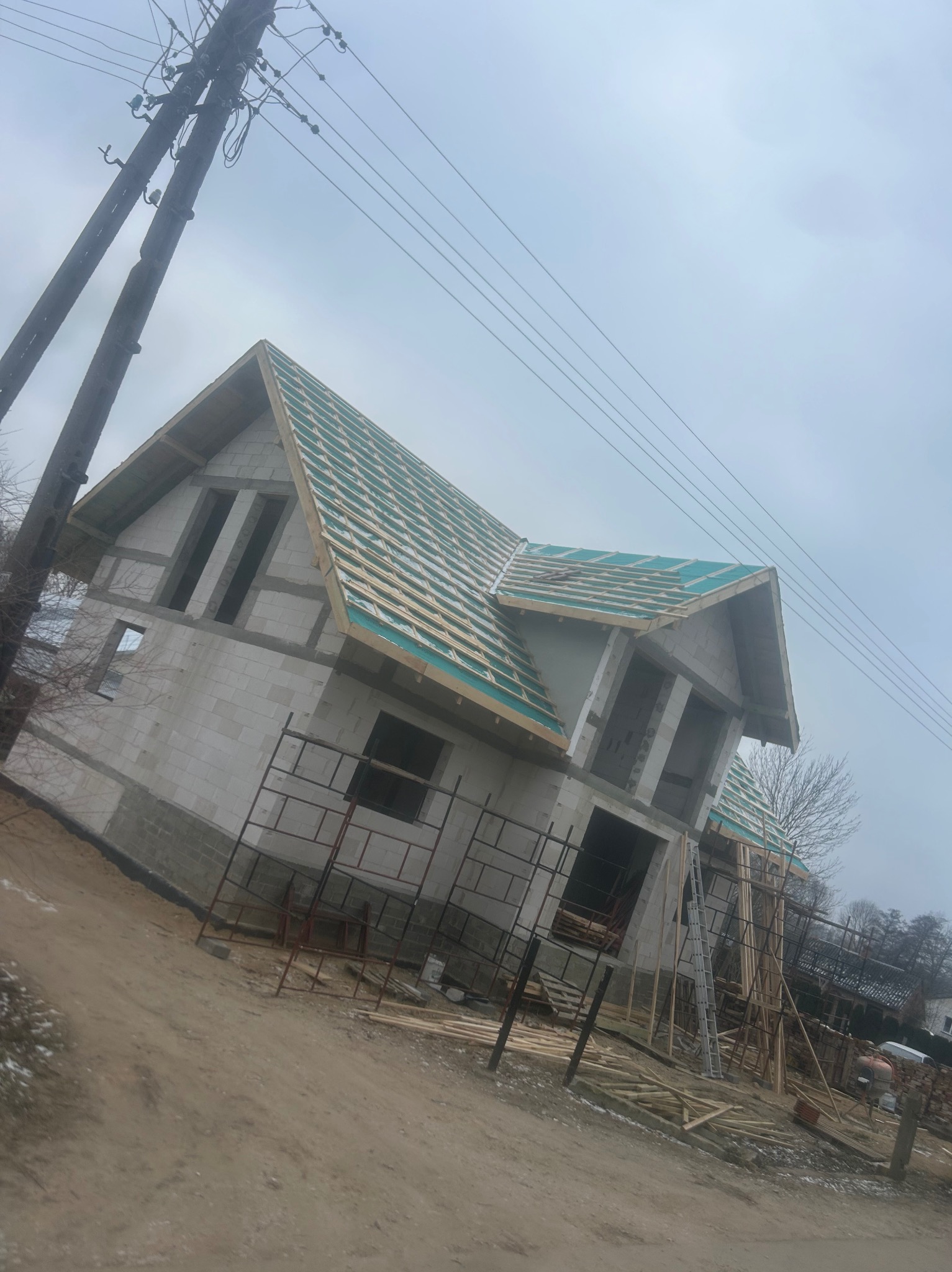 Surowy stan domu jednorodzinnego w trakcie budowy, widoczne elementy konstrukcji dachu z deskowaniem i folią, ściany z betonu komórkowego, rusztowania i materiały budowlane na placu budowy...