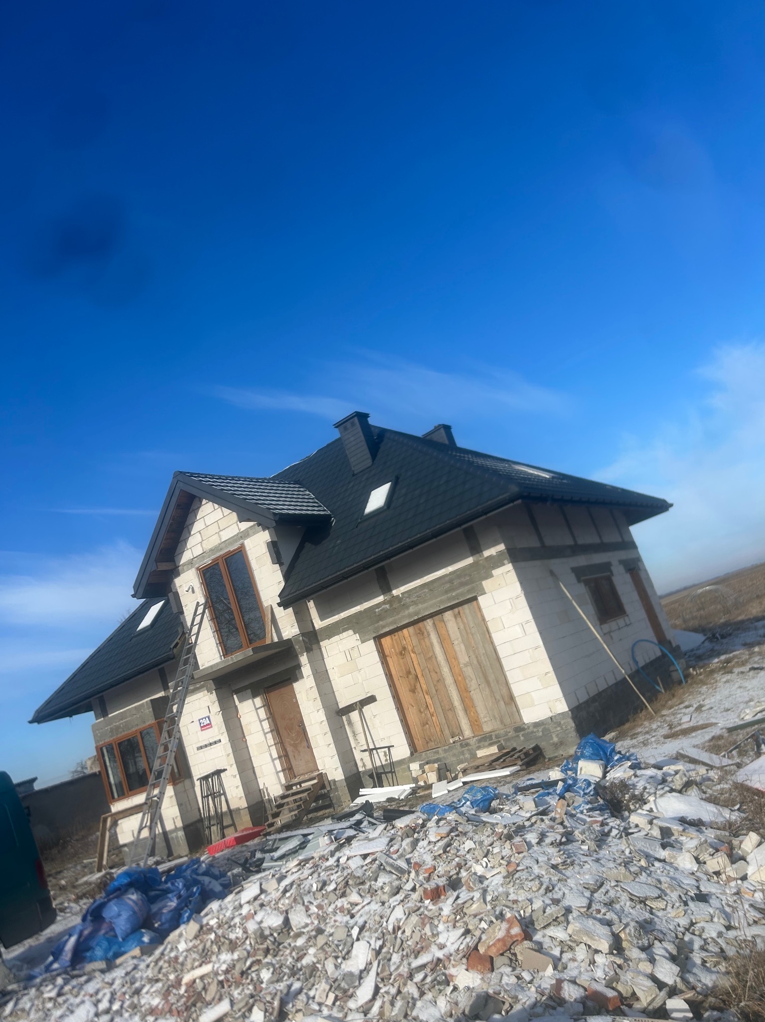 Dom w stanie surowym zamkniętym z czarnym dachem z dachówki bitumicznej, widoczne rusztowanie i gruz na posesji, błękitne niebo w tle.