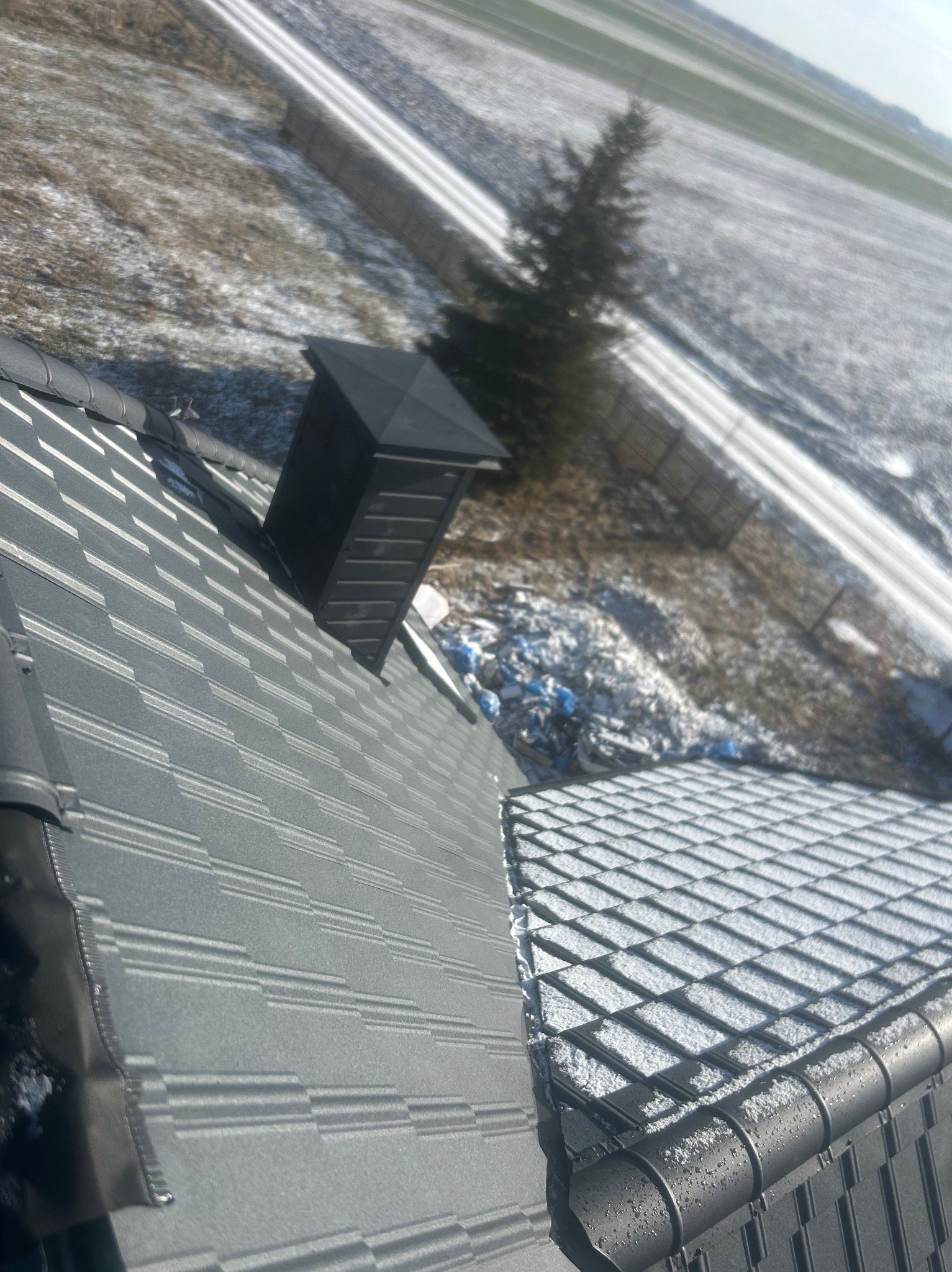 Szary dach z blachodachówki z kominem i rynną w zimowej scenerii, widok z góry na zaśnieżone pole i drzewo.