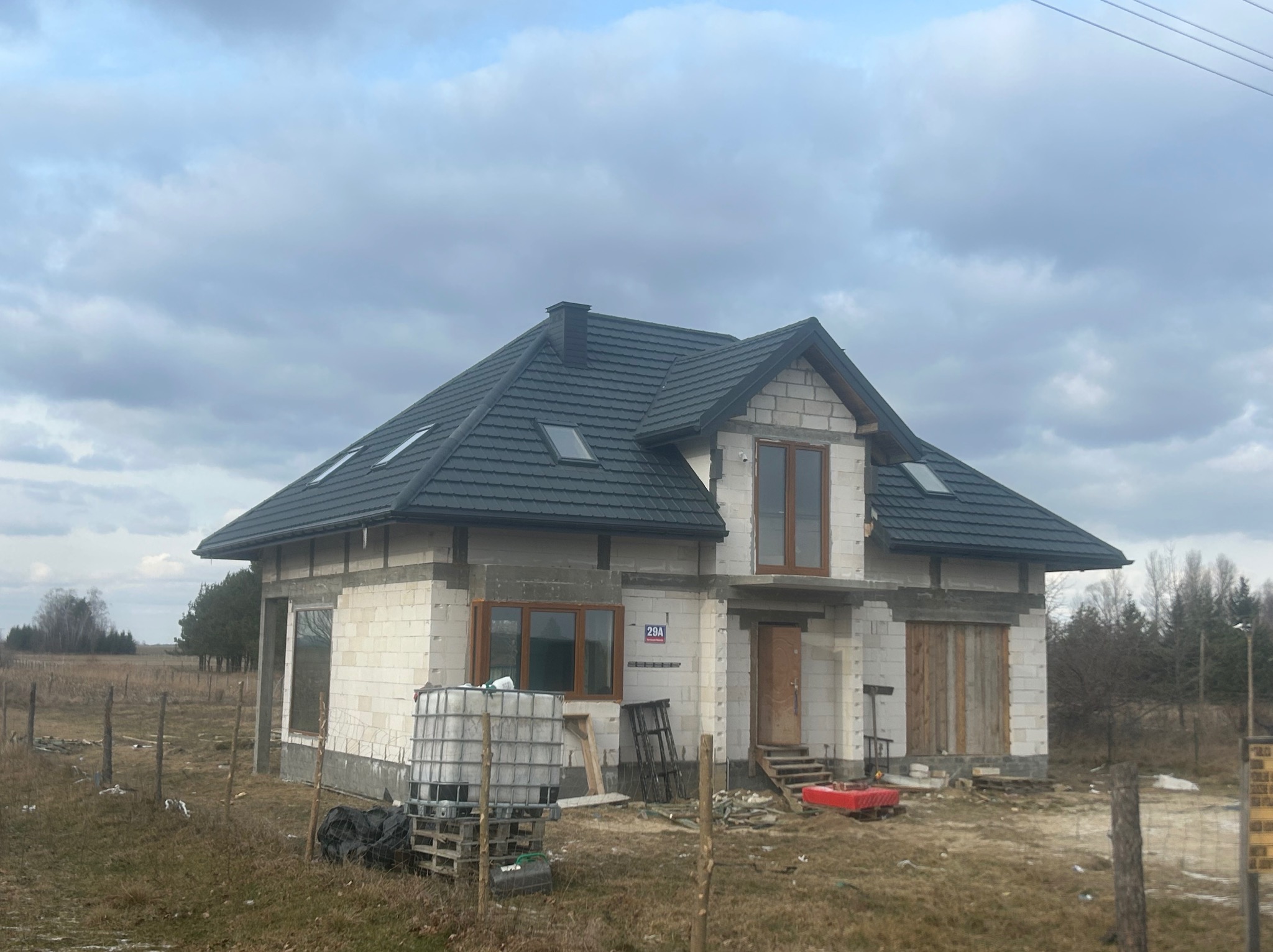 Dom jednorodzinny w trakcie budowy, z ciemnoszarą dachówką, widoczne okna dachowe, ściany z białego bloczka, numer domu 29A, teren wokół niezagospodarowany.
