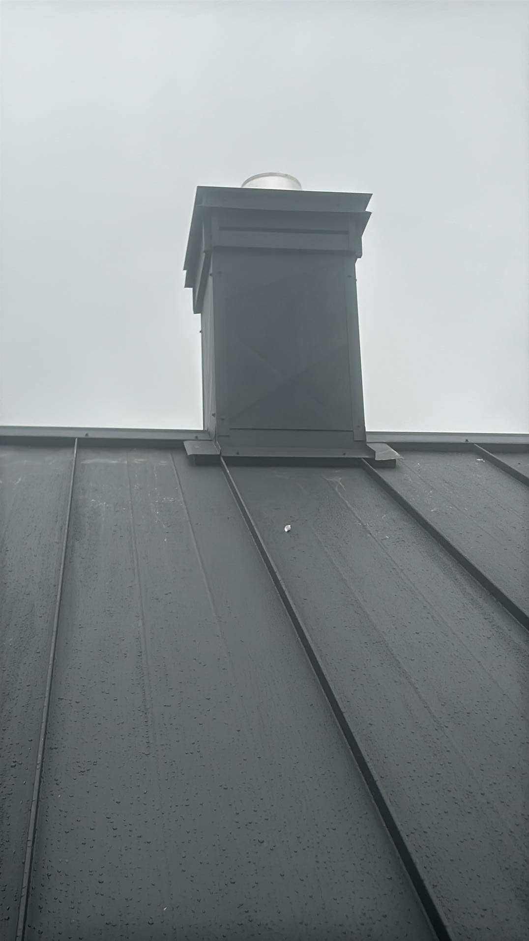 Szary metalowy dach z kominem w pochmurny dzień, widoczne krople deszczu.