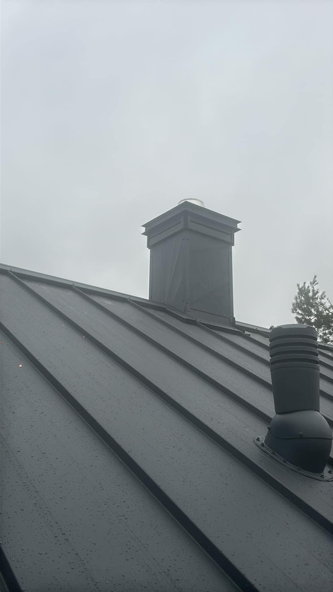 Metalowy dach z kominem i wentylacją w pochmurny dzień, widoczne krople deszczu.