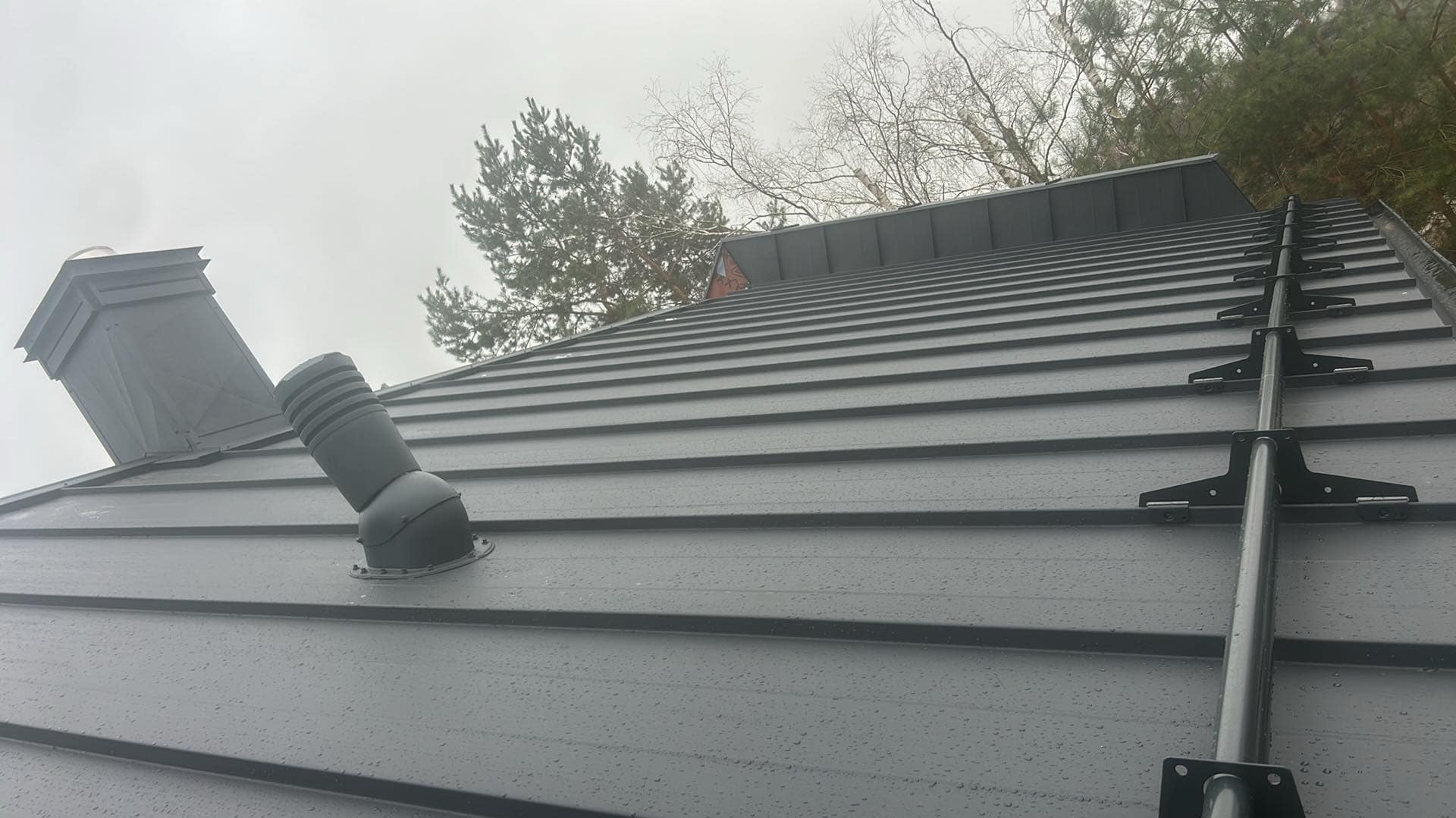 Szary dach z blachy na domu w Sterdyniu z kominem i wentylacją dachową, widoczna drabina dachowa. Widok w pochmurny dzień.