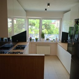 Mateusz Dębczyk - Nowoczesna kuchnia w Piotrkowie Trybunalskim z białymi, błyszczącymi szafkami górnymi i drewnopodobnym blatem, widok na okno z roślinami doniczkowymi.