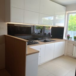 Mateusz Dębczyk - Nowoczesna kuchnia z białymi, błyszczącymi szafkami wiszącymi i dolnymi, drewniany blat, czarny panel szklany nad blatem, widok na okno z kwiatami, płytki podłogowe w odcieniach beżu.