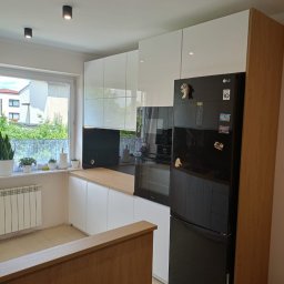 Mateusz Dębczyk - Nowoczesna kuchnia z białymi szafkami, drewnianym blatem, czarnym panelem nad blatem i czarną lodówką LG z magnesami, widok na okno z roślinami.