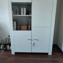 Mateusz Dębczyk - Biała szafa na wymiar z przeszkloną witryną, okrągłe kryształowe gałki, zabezpieczenie przed dziećmi na dolnych drzwiczkach, drewniana podłoga.