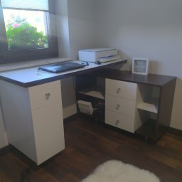 Mateusz Dębczyk - Narożne biurko z białymi frontami szuflad i ciemnobrązowym blatem, z laptopem i drukarką na blacie, widok na okno z roślinami w tle.