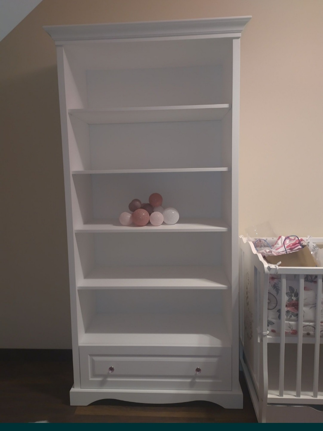 Biały regał z szufladą na dole, z kulami dekoracyjnymi na jednej z półek, stojący obok białego łóżeczka dziecięcego.