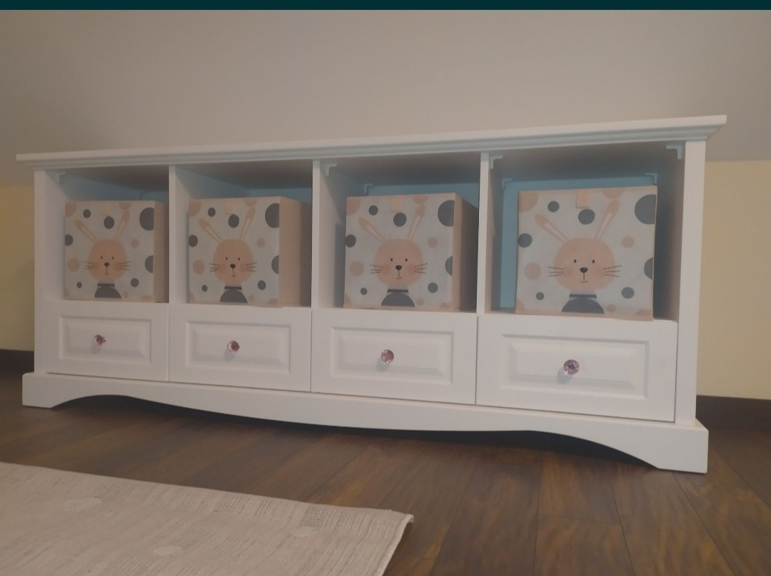 Biała szafka na zabawki z czterema szufladami i otwartymi półkami, w których umieszczono ozdobne pudełka z motywem królików, różowe kryształowe gałki, na tle drewnianej podłogi i fragmentu dywanu.