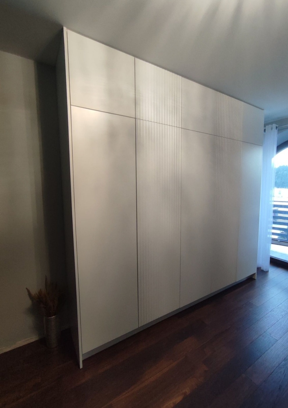 Duża, jasna szafa wnękowa z ryflowanymi frontami, widok z boku na tle drewnianej podłogi i okna z balkonem.