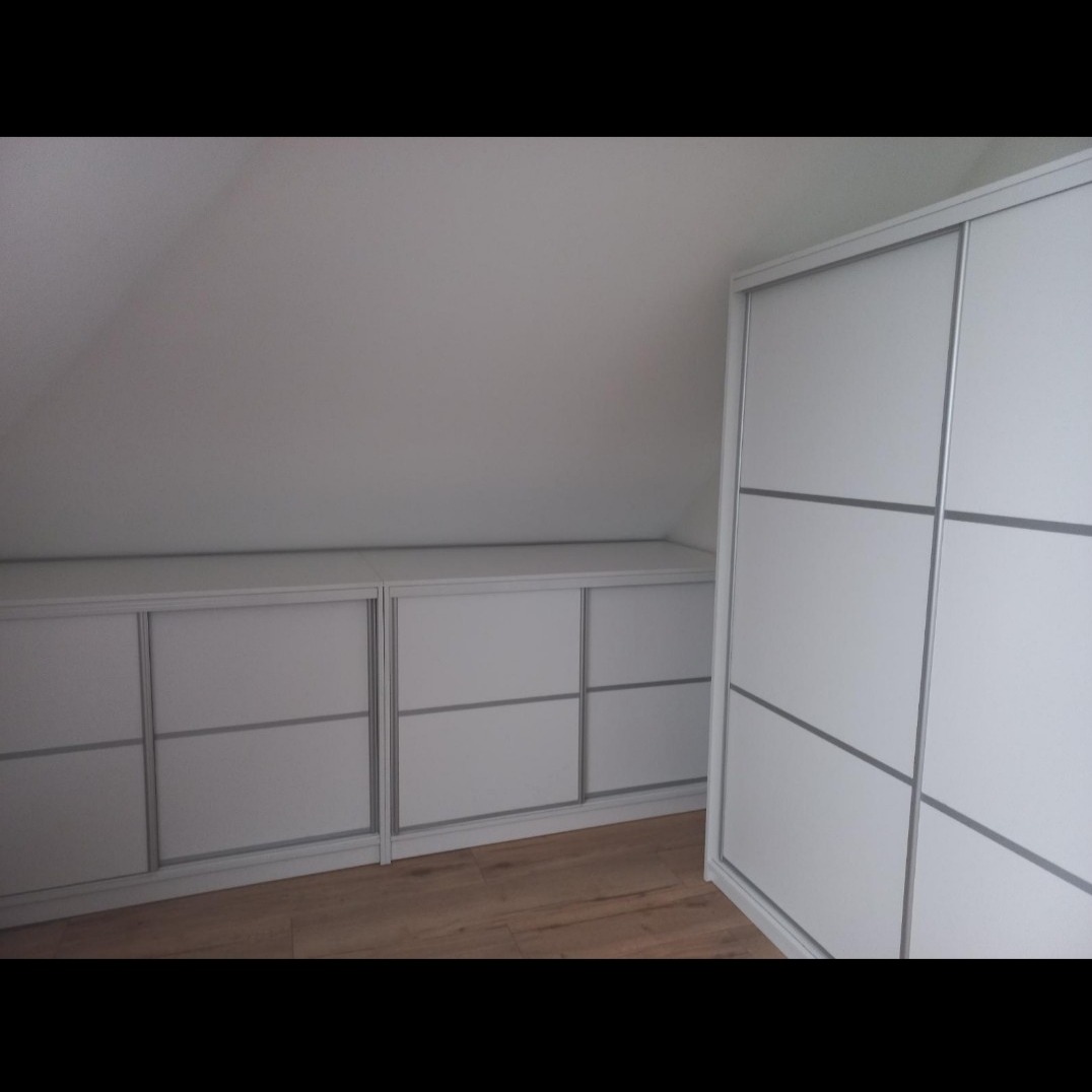 Biała zabudowa pod skosem z przesuwnymi drzwiami i szafa trzydrzwiowa z poziomymi podziałami, wykończona srebrnymi profilami.