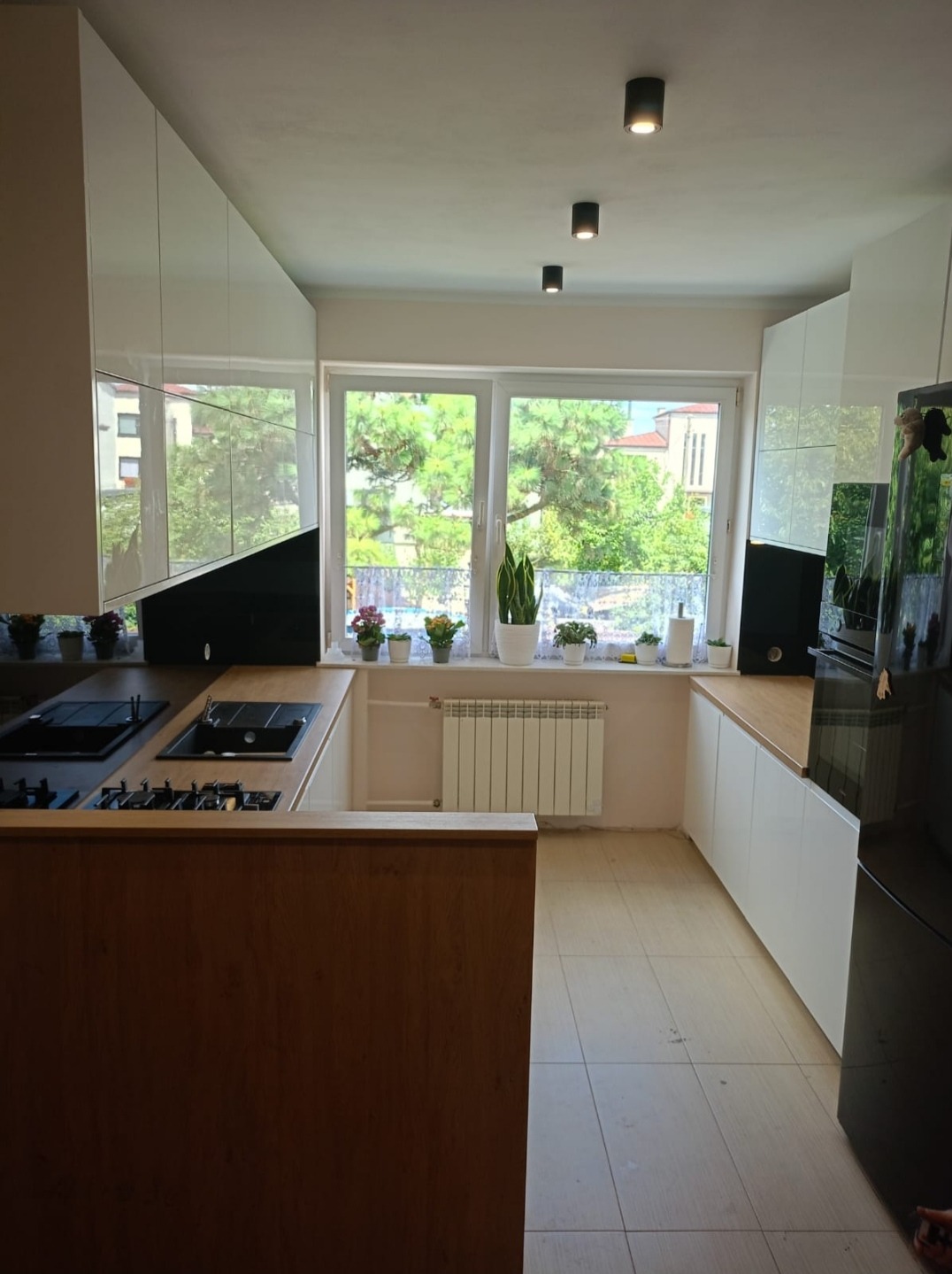 Nowoczesna kuchnia w Piotrkowie Trybunalskim z białymi, błyszczącymi szafkami górnymi i drewnopodobnym blatem, widok na okno z roślinami doniczkowymi.