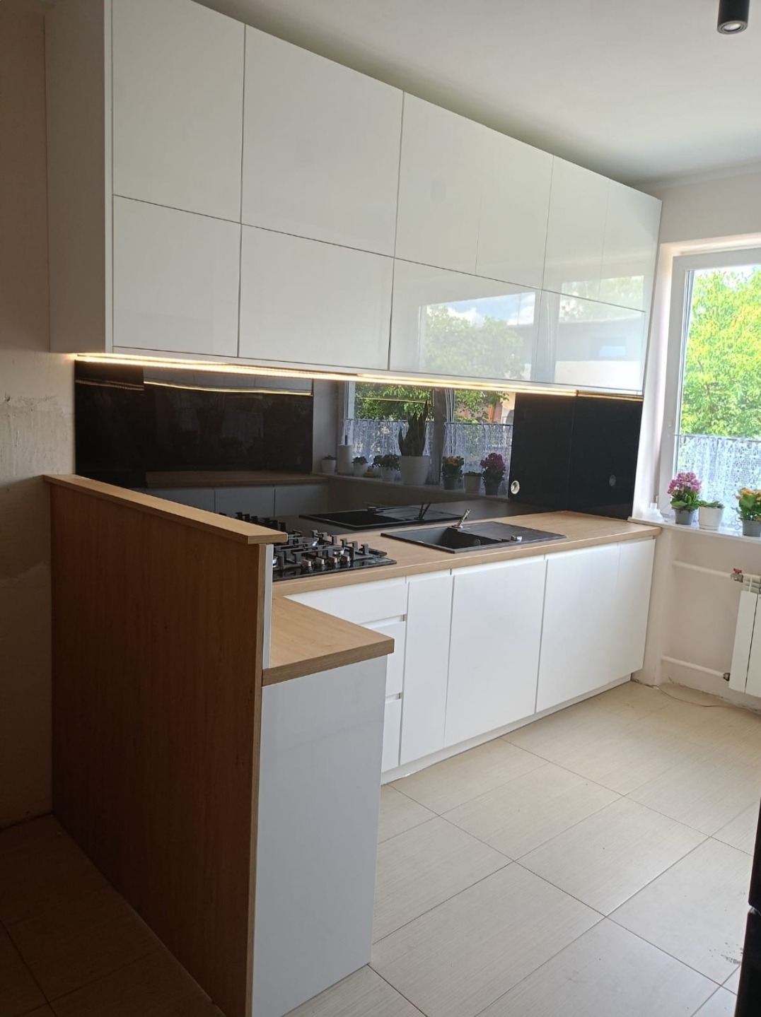 Nowoczesna kuchnia z białymi, błyszczącymi szafkami wiszącymi i dolnymi, drewniany blat, czarny panel szklany nad blatem, widok na okno z kwiatami, płytki podłogowe w odcieniach beżu.