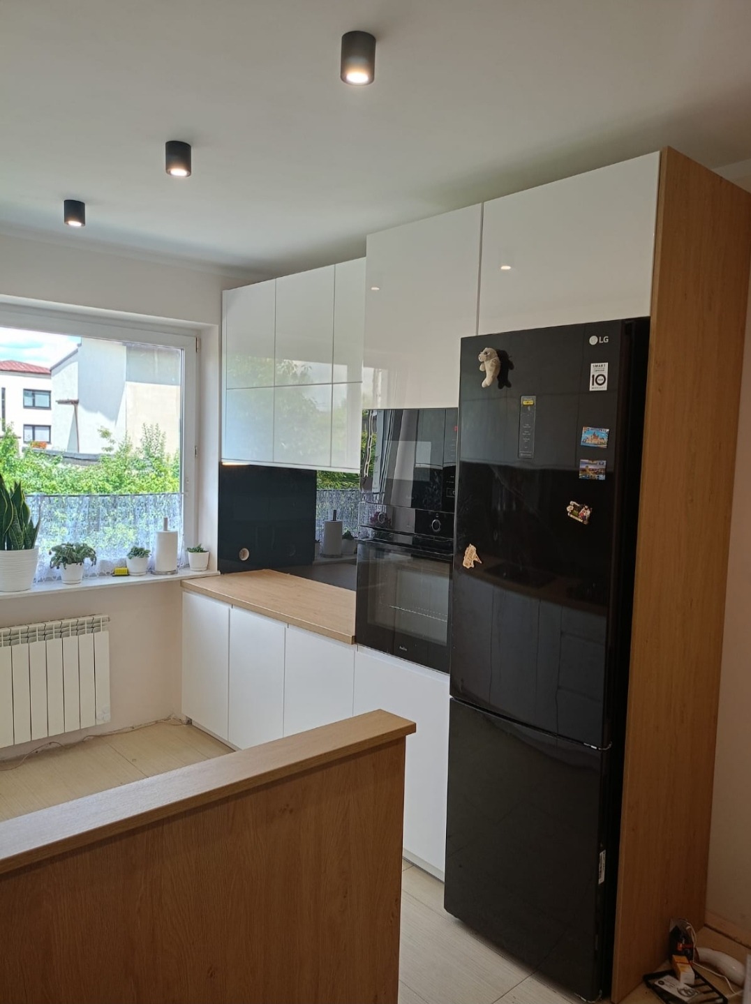 Nowoczesna kuchnia z białymi szafkami, drewnianym blatem, czarnym panelem nad blatem i czarną lodówką LG z magnesami, widok na okno z roślinami.