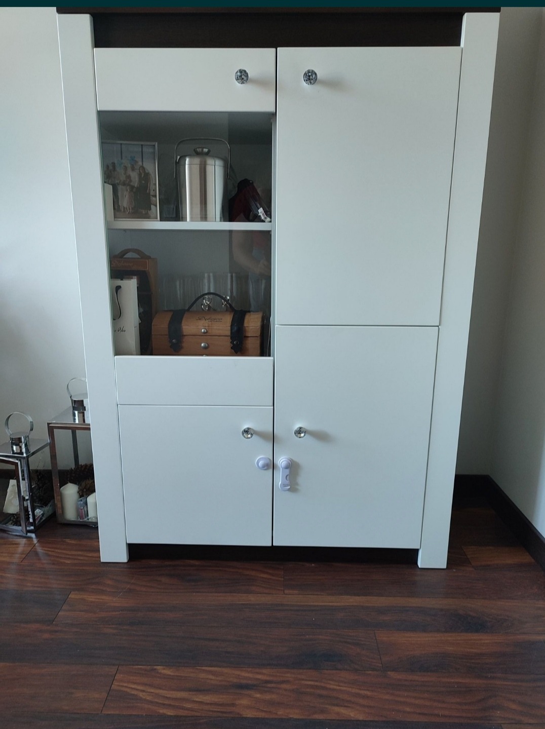 Biała szafa na wymiar z przeszkloną witryną, okrągłe kryształowe gałki, zabezpieczenie przed dziećmi na dolnych drzwiczkach, drewniana podłoga.